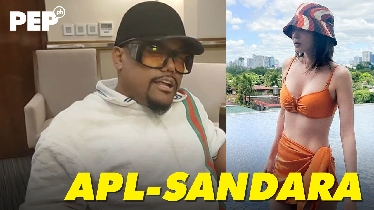 Apl.de.ap sitting wearing a cap and heavy-framed glasses. Sandara Parak in orange bathing suit.